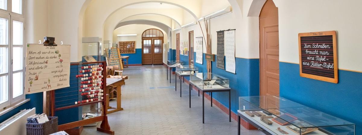 Dauerausstellung im ehemaligen Schulflur. Exponate im Schulflur, vorn das Thema Schreiben, links Fenster zum Schulhof.