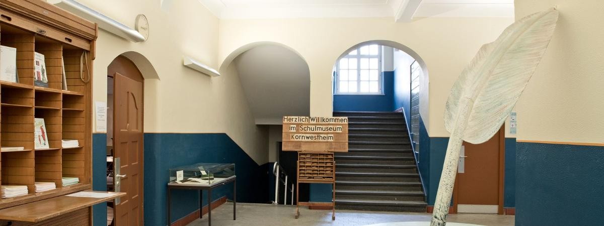Der Eingangsbereich mit großem Tintenfass, braunem Schrank, Vitrine, Hinweistafel und Treppenhaus im Hintergrund.