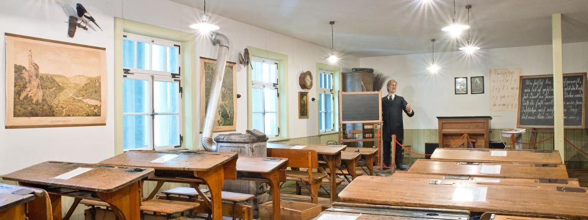 Blick ins historische Klassenzimmer mit Lehrerpuppe. Reihen historischer Schülerpulte, Tafel, Schulwandbilder, Lehrerpuppe.