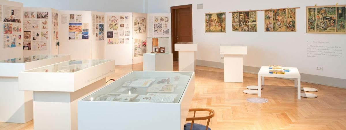 Blick in den Raum "Kindheit im Spiegel der Fibel". Weiße Vitrinen mit Fibeln und anderem, Textafeln, Schulwandbilder mit Märchenmotiven an weißen Wänden, Tür zum Flur.