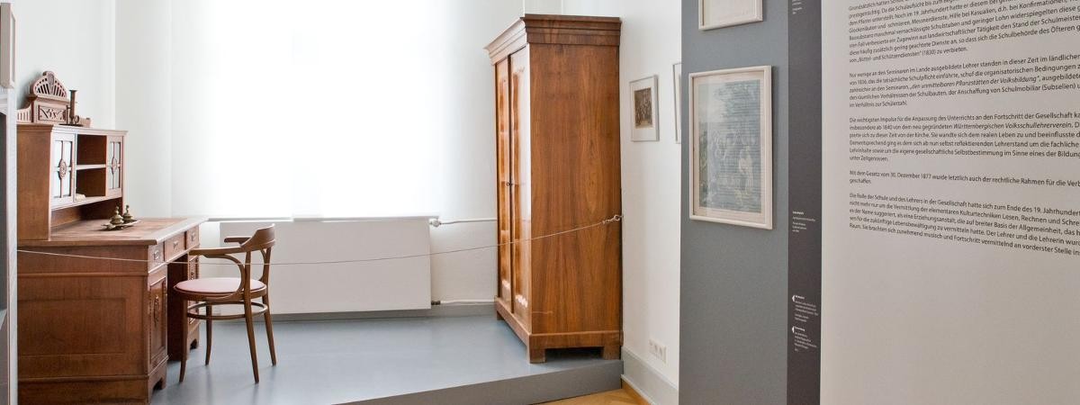 Blick in den Raum zum Thema "Lehrer". Standvitrinen mit Objekten, Texttafeln, dunkler Holzschreibtisch.