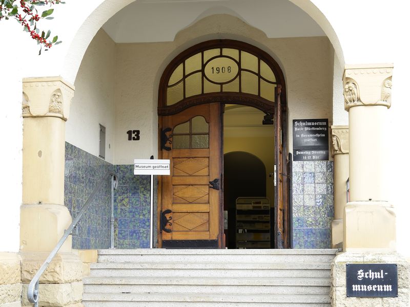 Eingangstür mit Treppe zum Schulmuseums, über der Tür ein Schild mit dem Baujahr 1913. Link zur Unterseite "Aktuelles".